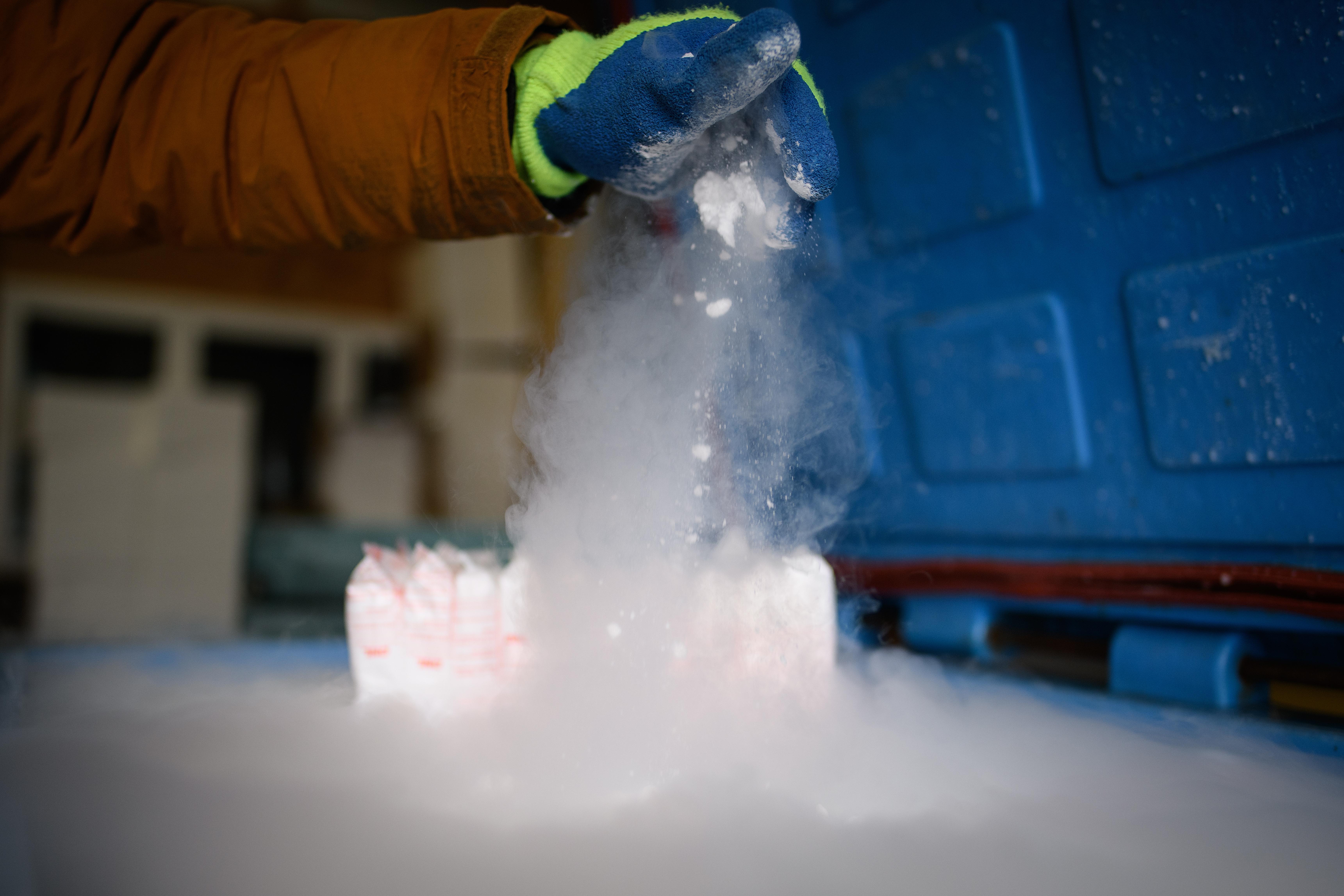 Someone wearing a thick blue glove drops dry ice on a surface at the bottom of the frame, causing white fog to spread 