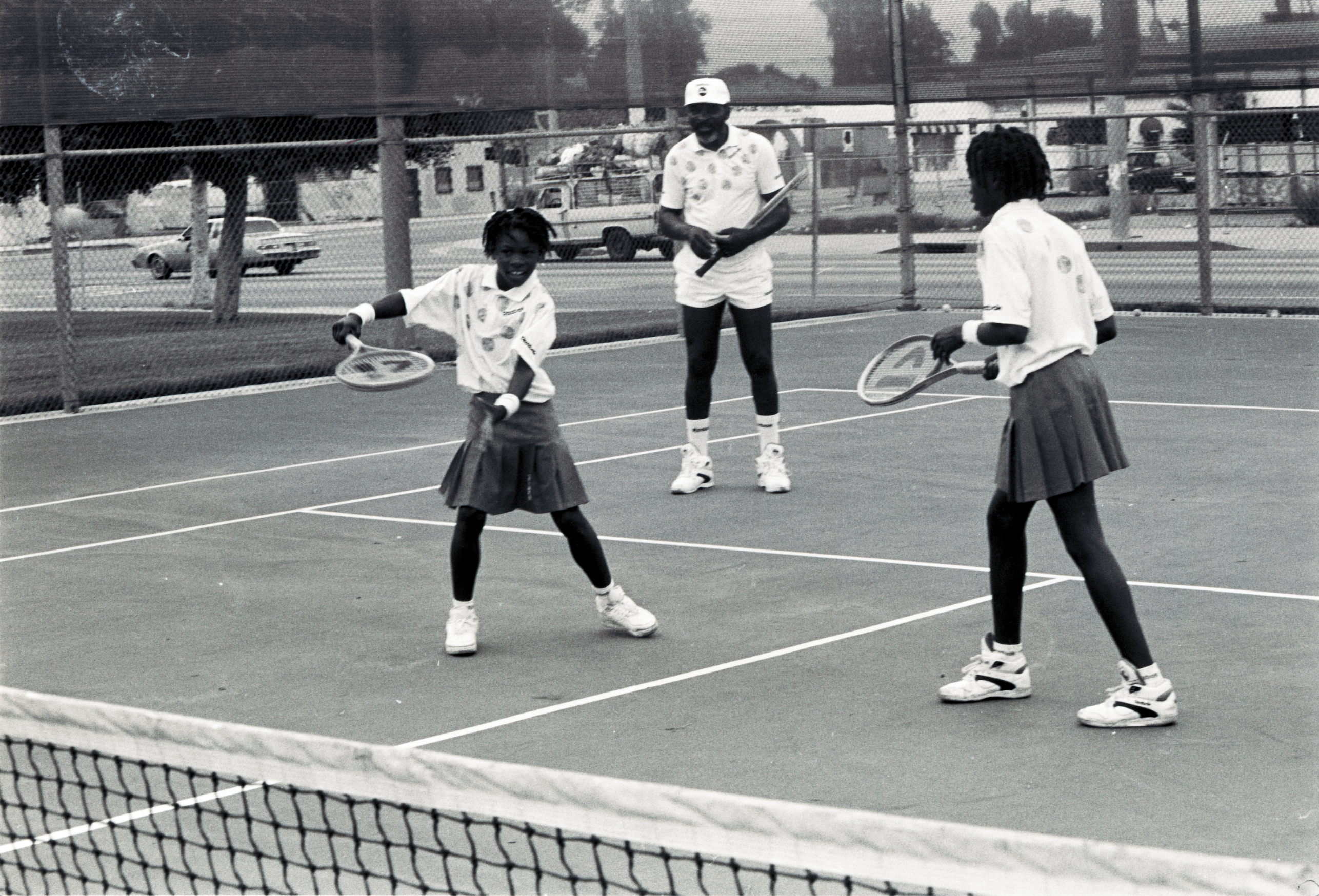 The sisters stand in front of their father, Richard Williams, who appears on the court behind them.