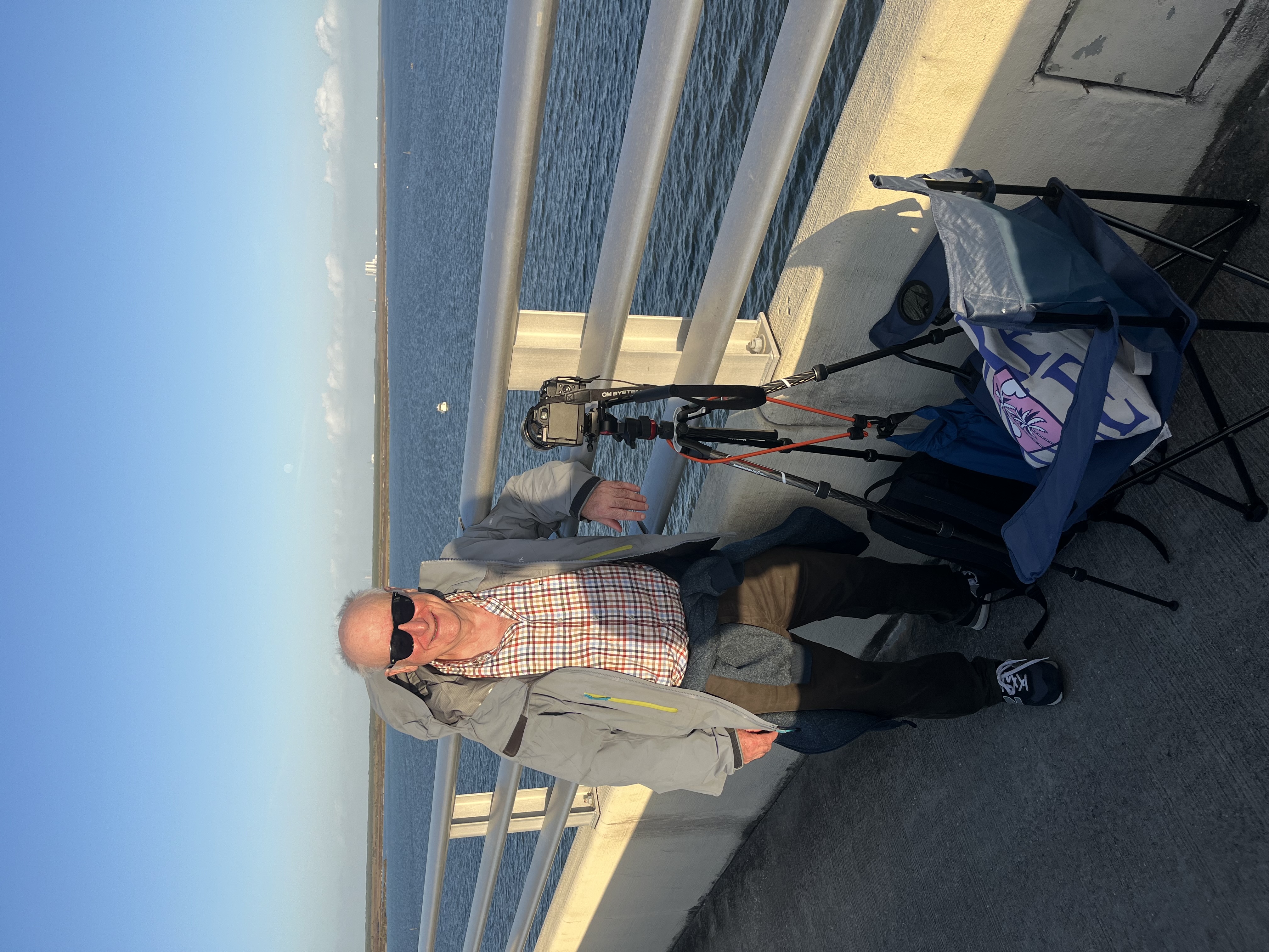 a man in sunglasses and a jacket stands near the side of a bridge with a camera and folding chair in front of him