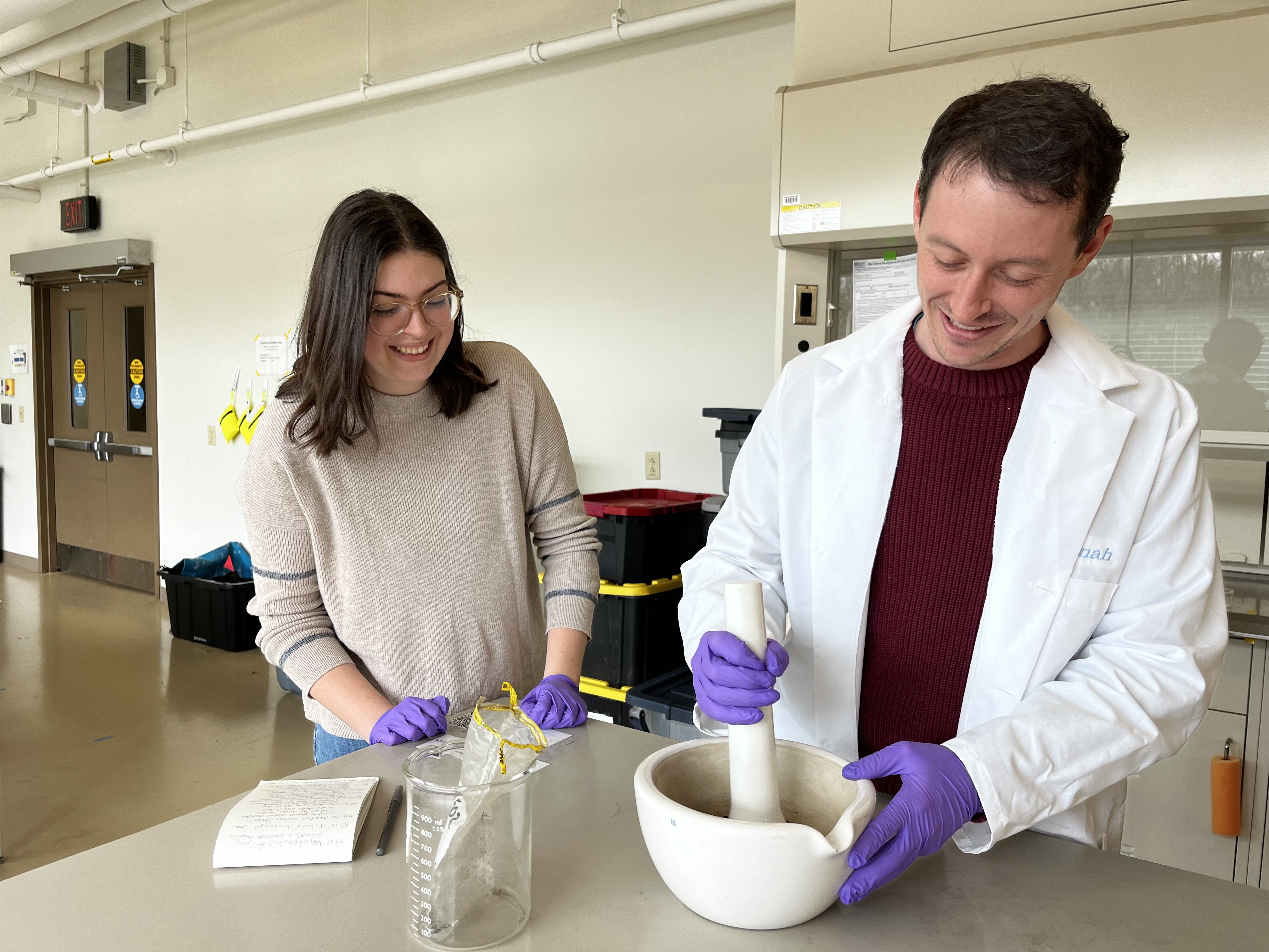 Two scientists in purple gloves stand beside a lab table. One is grinding a sample in a white bowl while the other looks on and smiles.