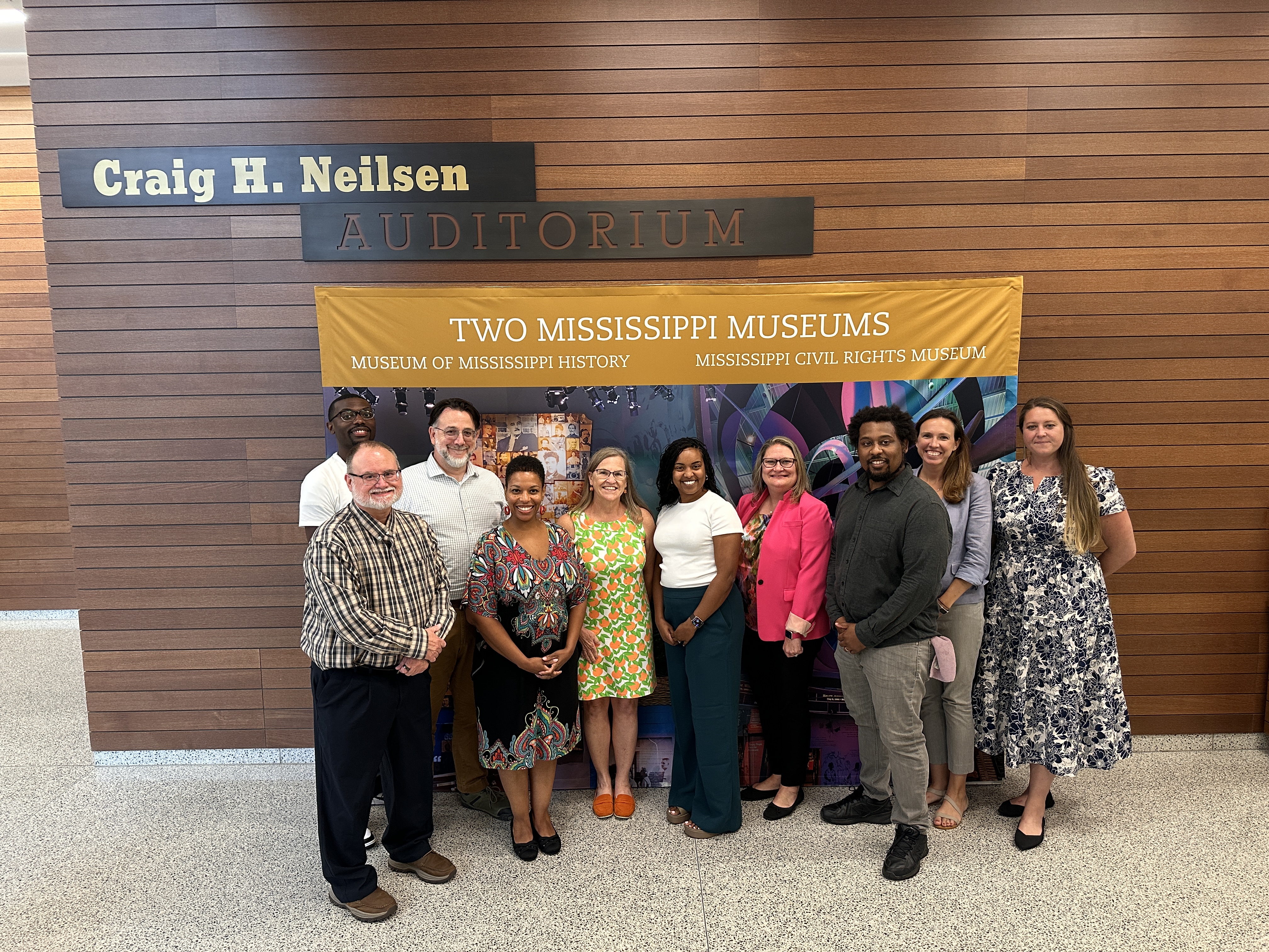 10 museum and education professionals dressed in business attire smiling and standing in front of a multicolored backdrop that reads, “Two Mississippi Museums, Museum of Mississippi History, Mississippi Civil Rights Museum”.
