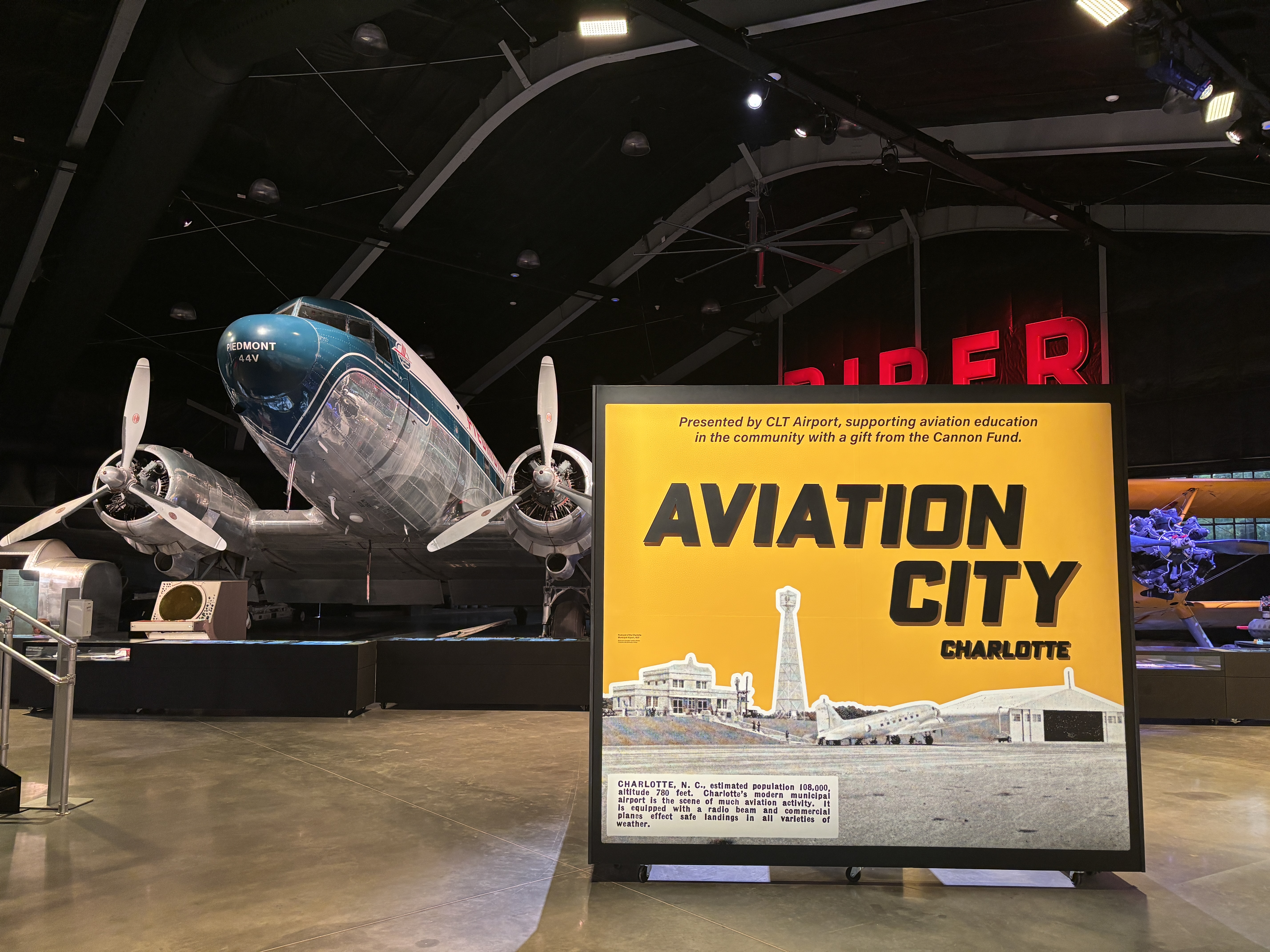 A propellor plane sits inside a hangar with a sign reading Aviation City in the foreground.